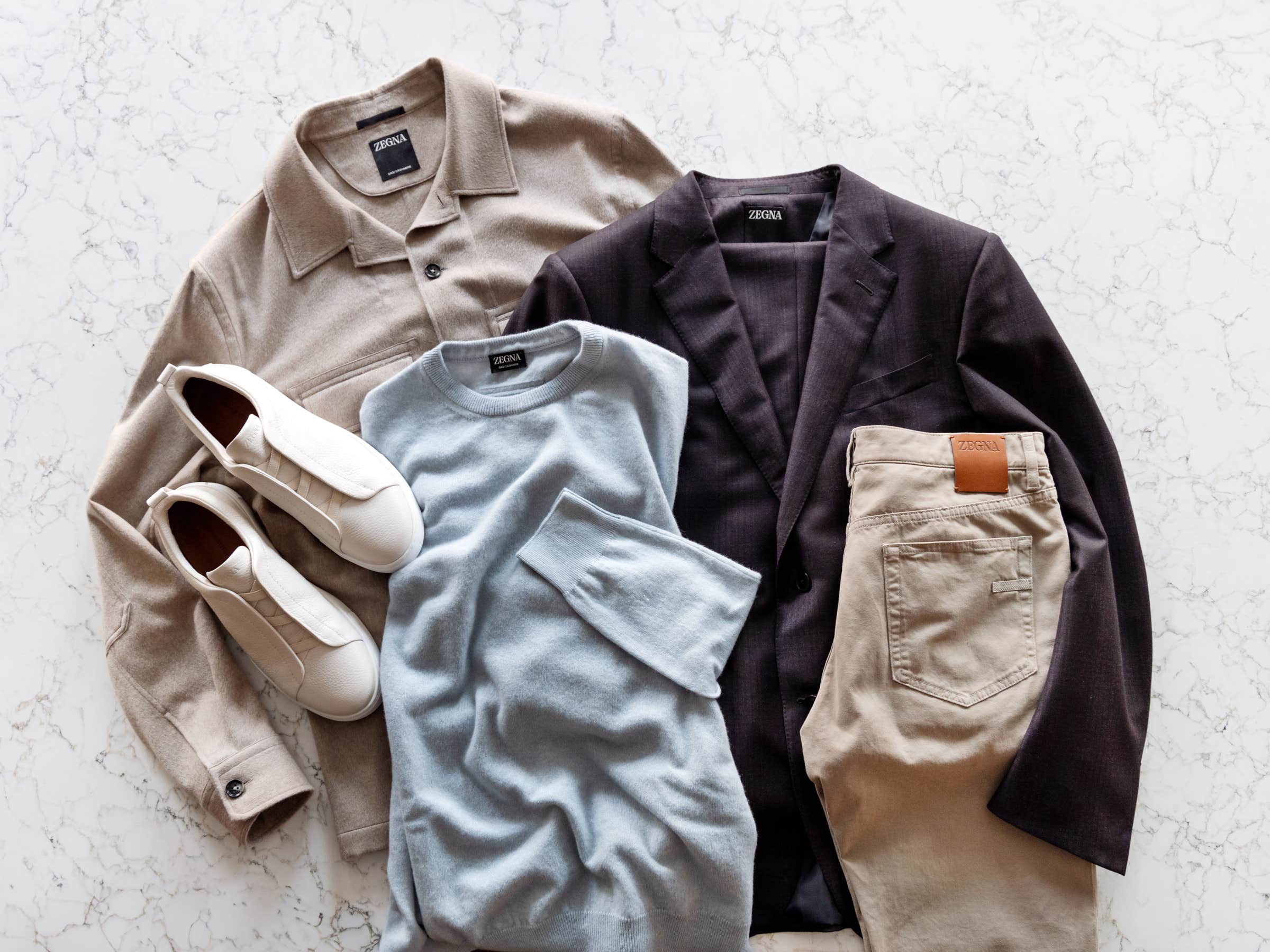 ZEGNA Oasi Cashmere Overshirt, ZEGNA Oasi Cashmere Crewneck, ZEGNA Trofeo Wool Burgundy Suit, ZEGNA Khaki City Fit 5-Pocket Jean and ZEGNA Triple Stitch Sneaker all laid out on a marble table